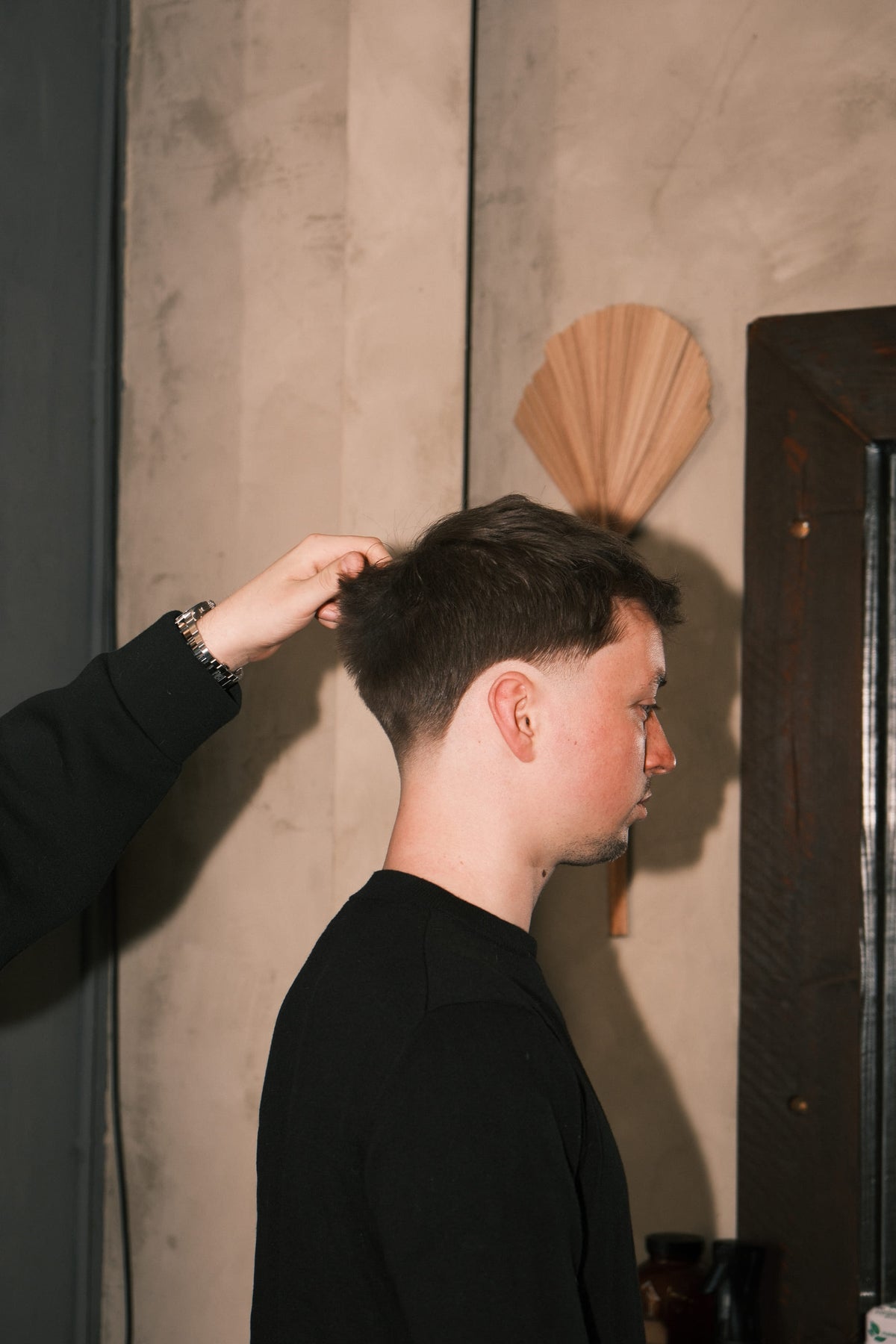 Klant in profiel, hand tilt het haar bij de kruin; betonnen wand met gedroogd palmblad.