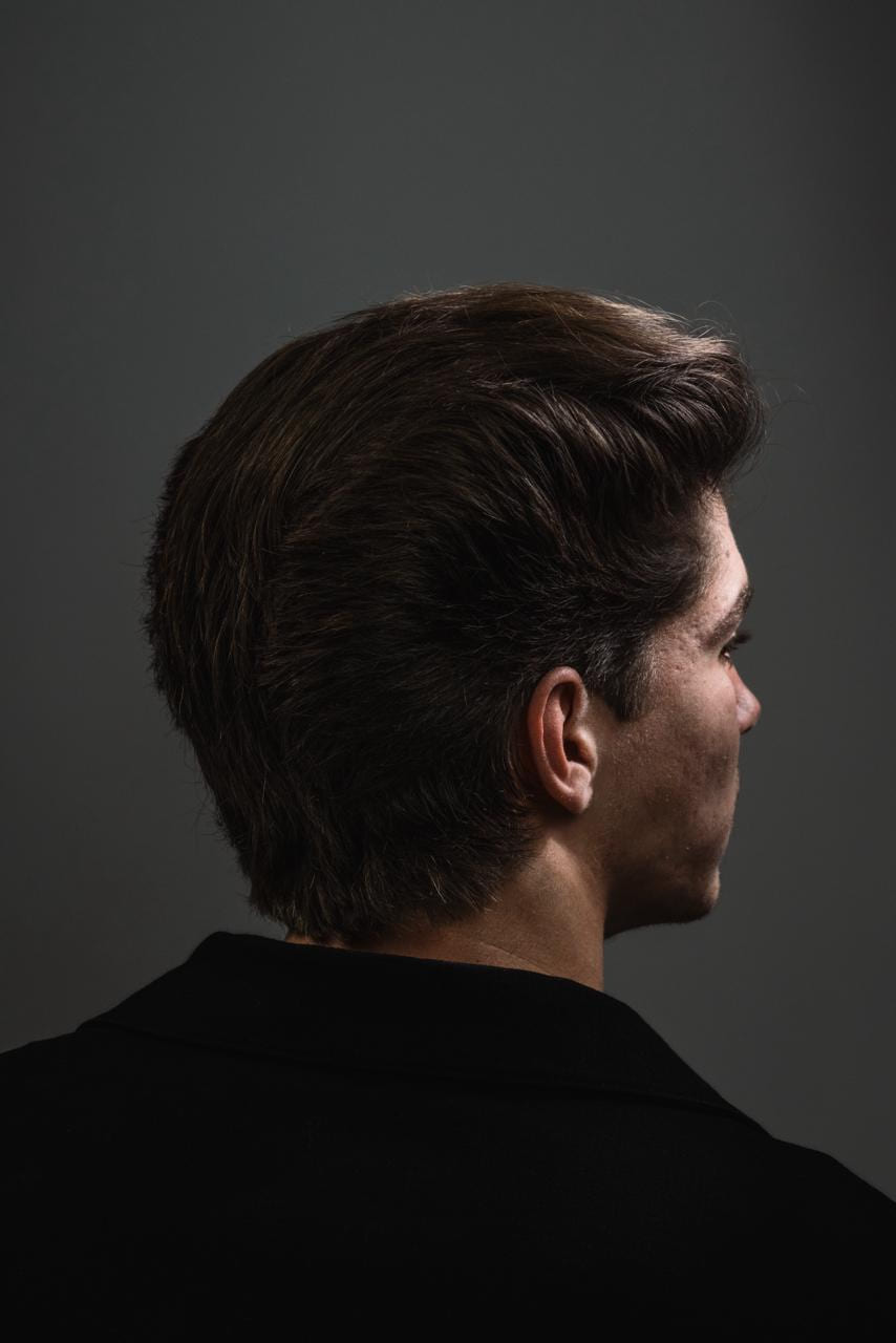 Achterhoofd en profiel van een volumineuze naar achteren gekamde coupe; donkergrijze studiogrond, zwart poloshirt.