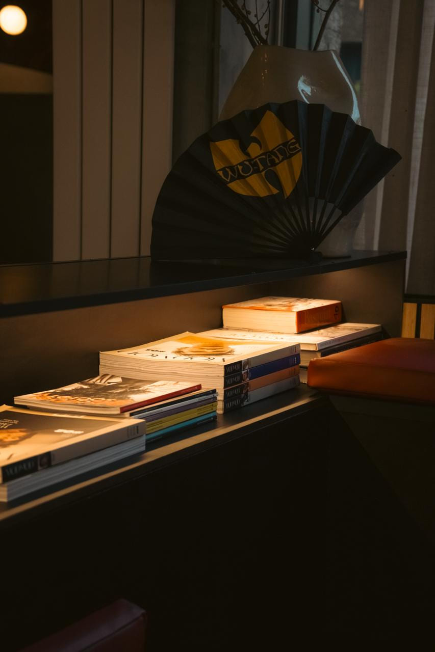 Stapels boeken en tijdschriften op de bank, met een zwart-gele Wu-Tang-waaier tegen de vaas.
