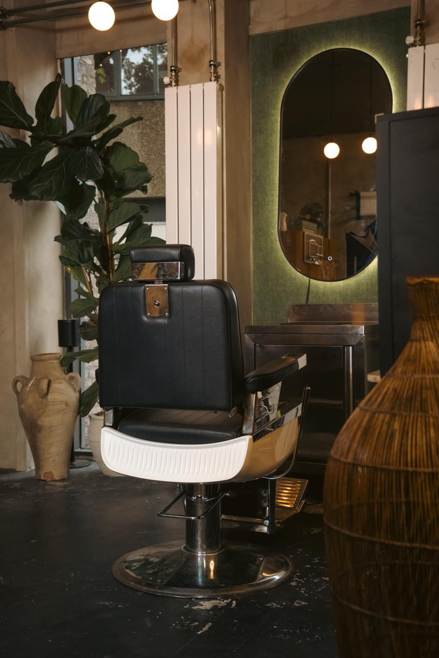 Klassieke barberstoel voor een groen paneel met verlichte ovale spiegel, vioolbladplant en terracotta kruik ernaast.
