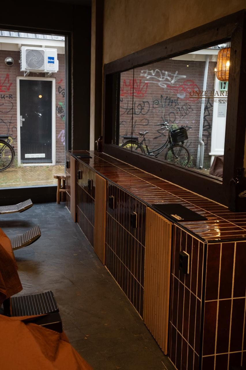 Rij barberstoelen langs de tegelwerkbank, raam met beklade muur en fietsen erachter.