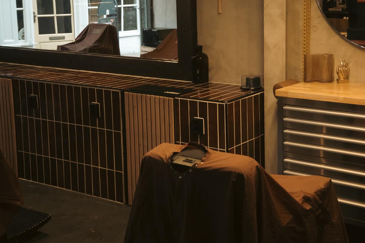 De volle breedte van de werkbank: bruine stroktegels, spiegelwand, stoelen onder hoezen, gereedschapskast met houten blad.