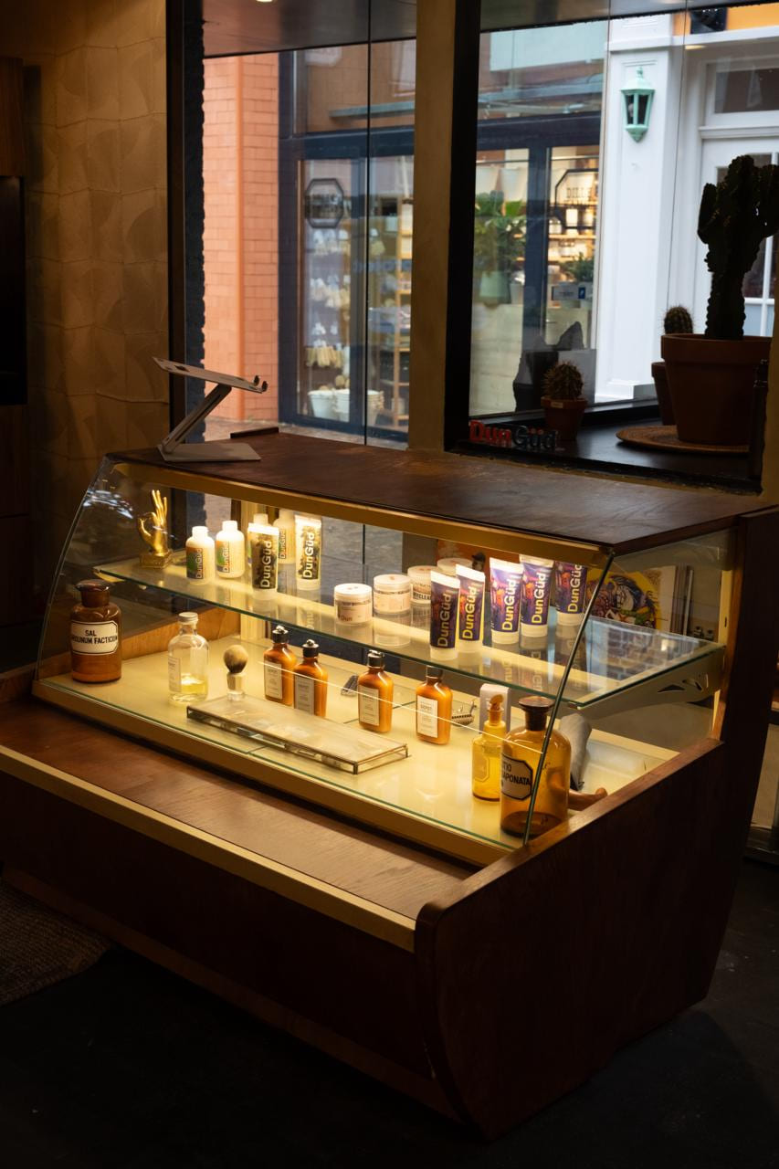 De gebogen glazen vitrine schuin van voren: DunGüd-tubes op de bovenplank, apothekersflessen en Depot-flacons eronder.